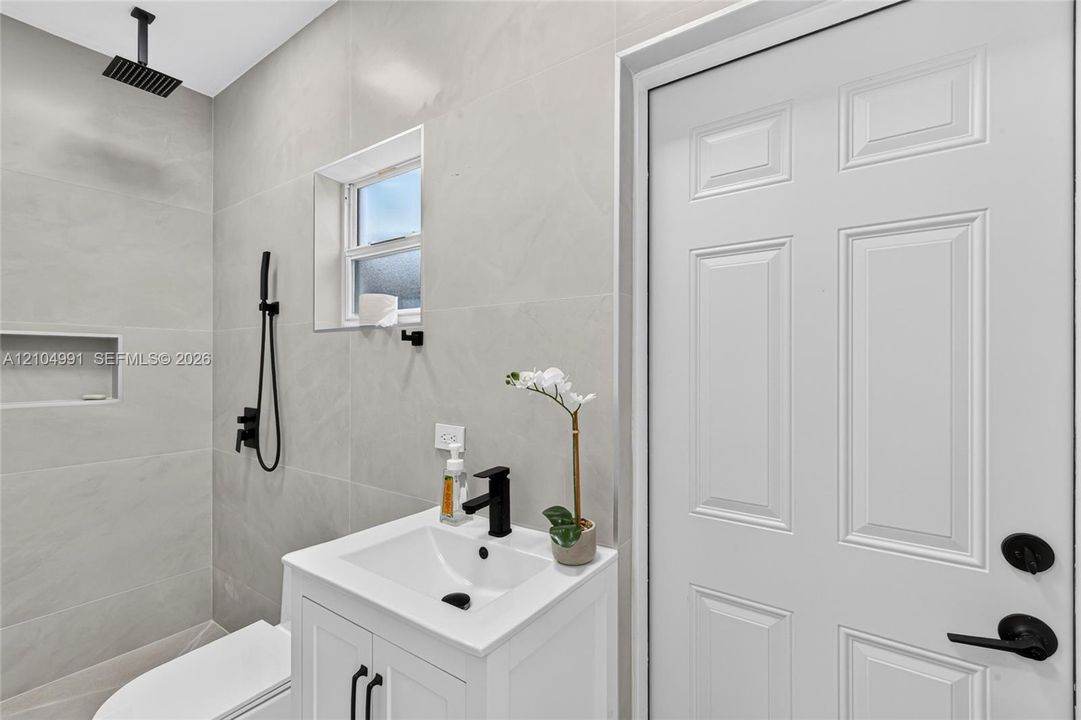 Full-height neutral wall tiling, walk-in shower with rainfall showerhead and built-in wall niche, white vanity with rectangular basin and matte black fixtures, and six-panel interior door with matte black hardware