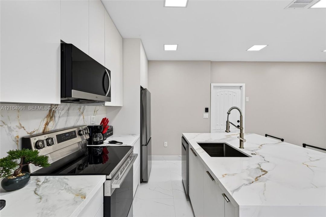 Kitchen featuring white slab cabinetry, large-format tiled flooring, stone-finish countertops with matching full-height backsplash, stainless steel appliances, and a central island with a high-arc faucet