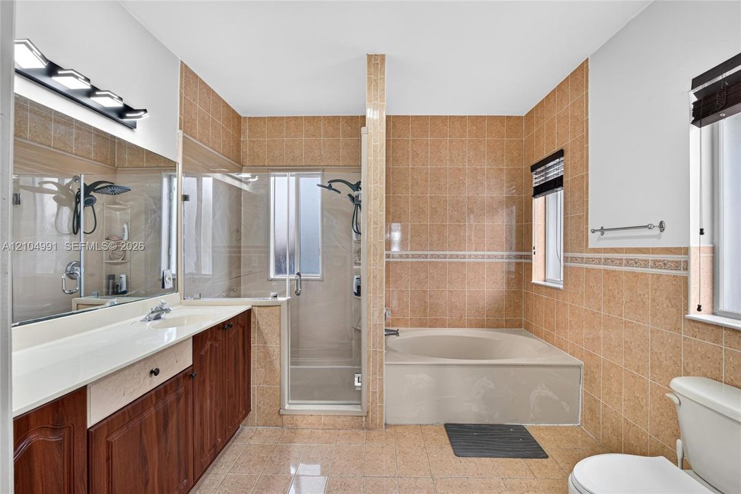 Bathroom featuring a dual-sink wood-finish vanity with an integrated countertop, a glass-enclosed shower, a deep soaking tub, and tile wall surrounds