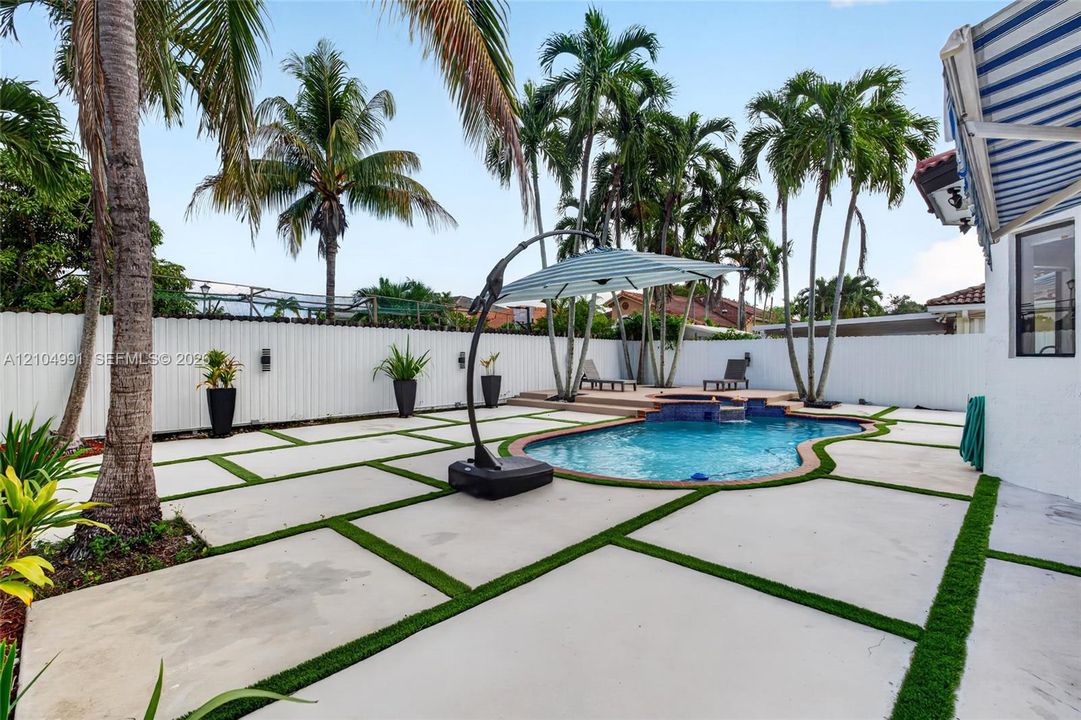 Outdoor living area featuring a swimming pool with a raised spa, concrete paver patio with turf inlays, privacy fencing, and mature palm trees