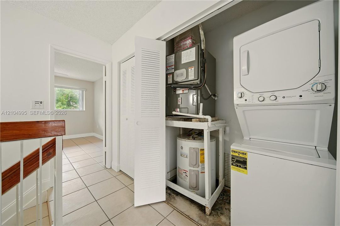 Utility closet containing a water heater and HVAC unit, accompanied by a white stacked washer and dryer, finished with tiled flooring