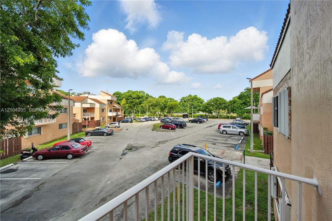 Elevated balcony with a white metal railing overlooking a paved parking area and mature trees
