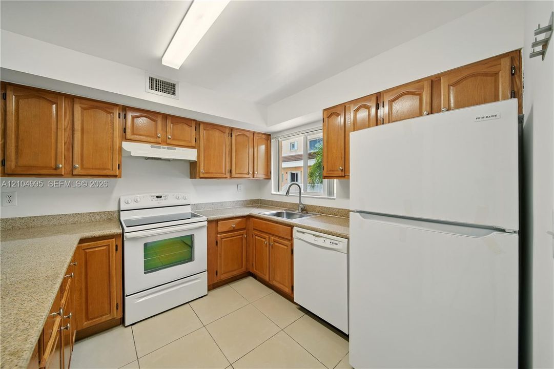 Kitchen featuring wood cabinetry, stone-finish countertops, tile flooring, and white appliances including a range, dishwasher, and refrigerator