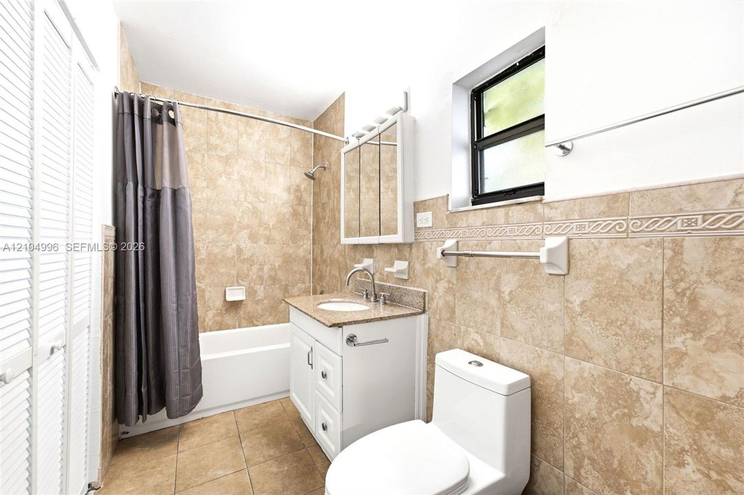Bathroom featuring neutral-toned stone-finish wall and floor tiling with decorative border inlay, white vanity with integrated stone-finish countertop, undermount sink, and mirrored medicine cabinet