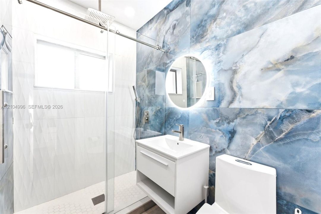 Large-format blue stone-finish wall tile, frameless glass shower enclosure with rain-style showerhead, floating vanity, circular backlit mirror, and white dual-flush toilet