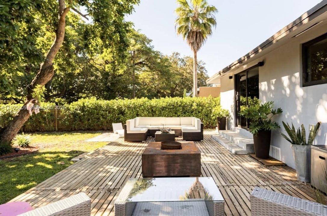 Expansive backyard featuring a wood-slat patio, a manicured lawn, lush perimeter hedging, and a white stucco exterior with dark-framed sliding glass doors