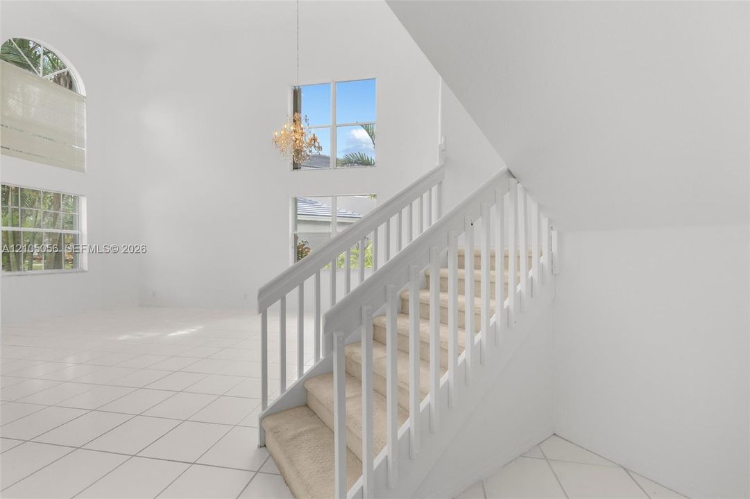 High-ceilinged social areas featuring a staircase with a white wooden railing, tile flooring, multiple windows, and a decorative hanging light fixture leading to upstairs landing
