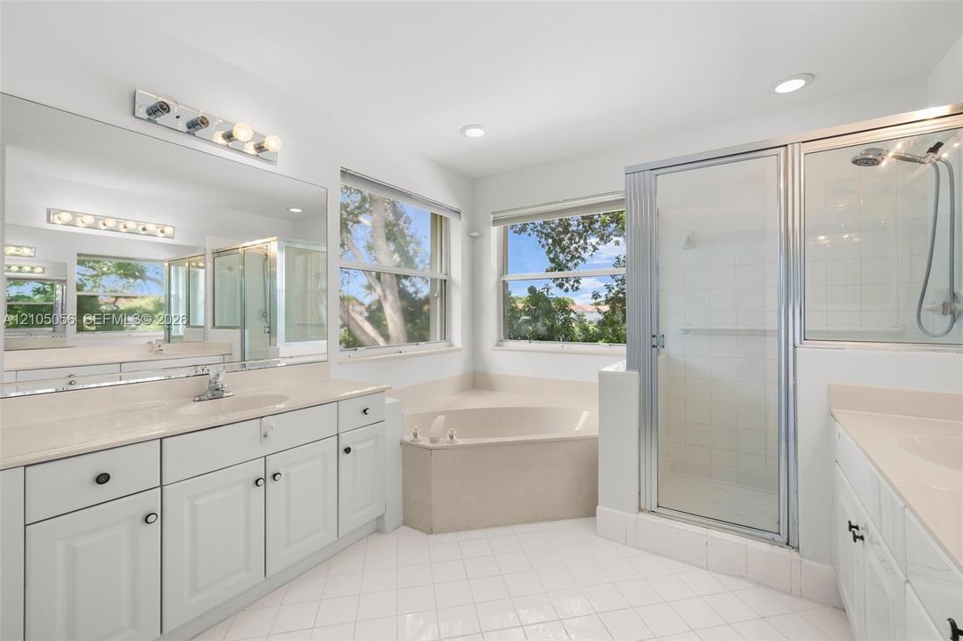 Primary bathroom featuring dual white cabinetry with light countertops, a corner soaking tub, a tiled shower stall with a metal-framed glass door, and large-format tile flooring