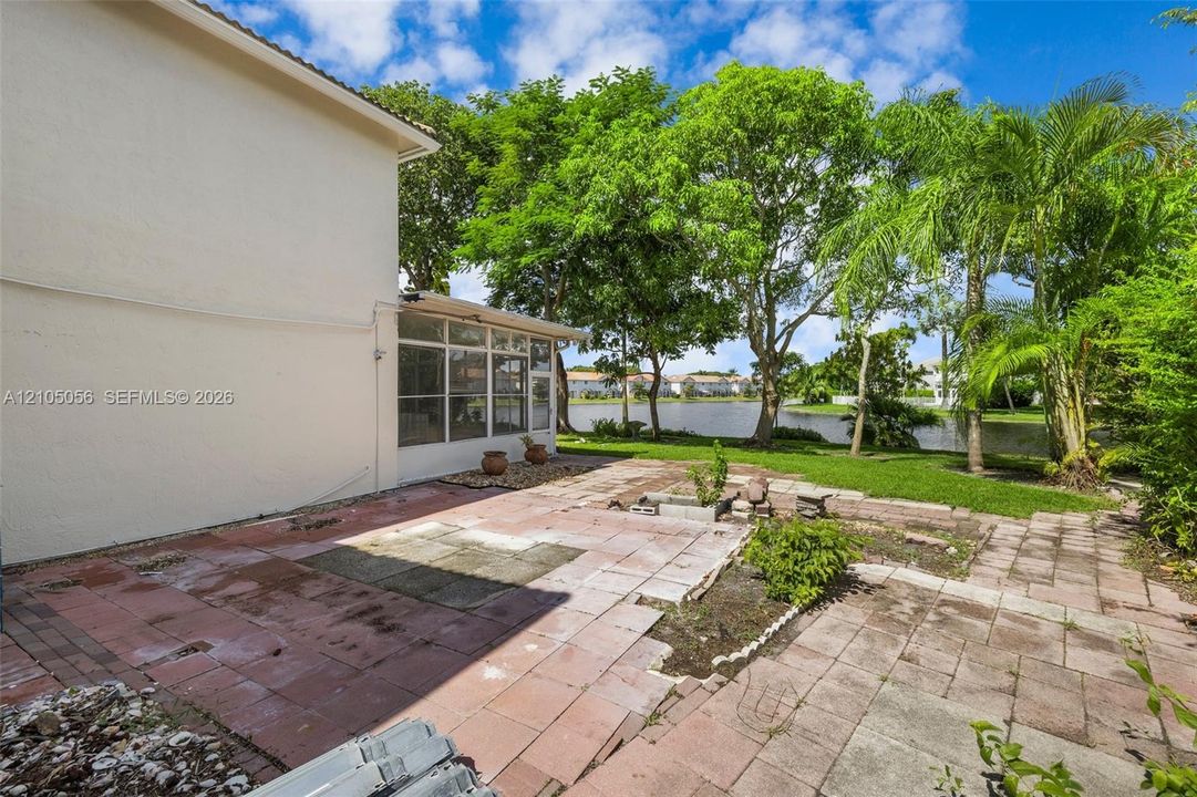 Rear exterior featuring a paver patio, screened-in enclosure, and waterfront grounds with mature trees and palm vegetation