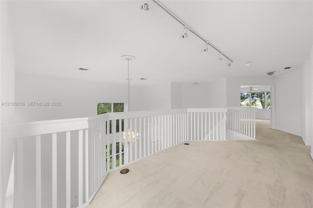 Open loft area featuring white vertical baluster railing, track lighting, a central pendant fixture, and neutral wall-to-wall carpeting leading to primary bedroom