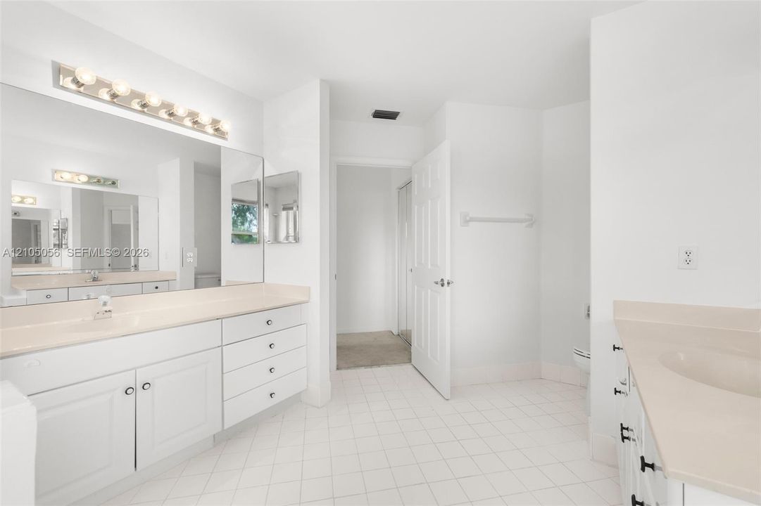 Dual vanity area featuring white cabinetry, solid-surface countertops, integrated sinks, full-width mirrors, and multi-bulb light fixtures over tiled flooring