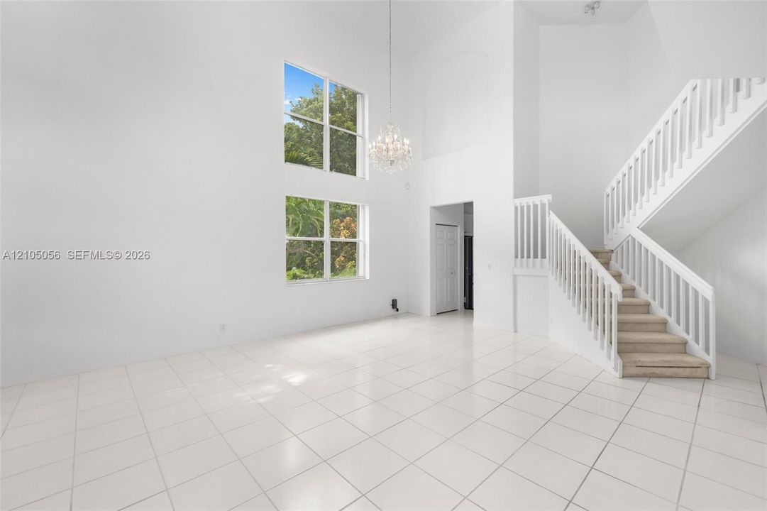 As you enter greeted by - Living Double-height ceiling, stacked windows, central crystal chandelier, white tile flooring, and staircase with white railings - Living room view to dining.
