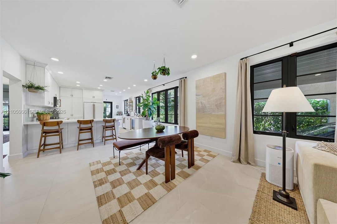 Open-concept kitchen and dining area featuring white shaker-style cabinetry, a central island with bar seating, large-format tile flooring, and oversized windows framed in black trim