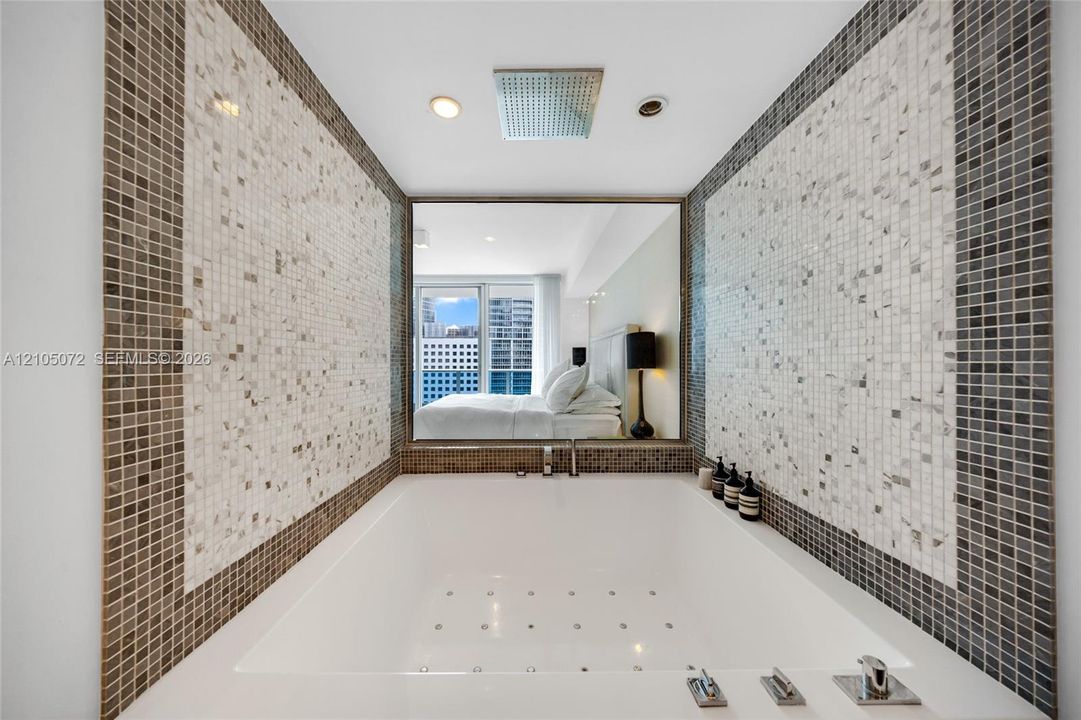 Built-in bathtub framed by mosaic tile and an interior window overlooking the primary bedroom.
