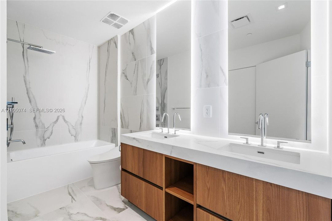 Dual-sink vanity with wood-finish cabinetry, quartz countertops, large-format tiled wall surround, undermount tub, and integrated LED mirror lighting