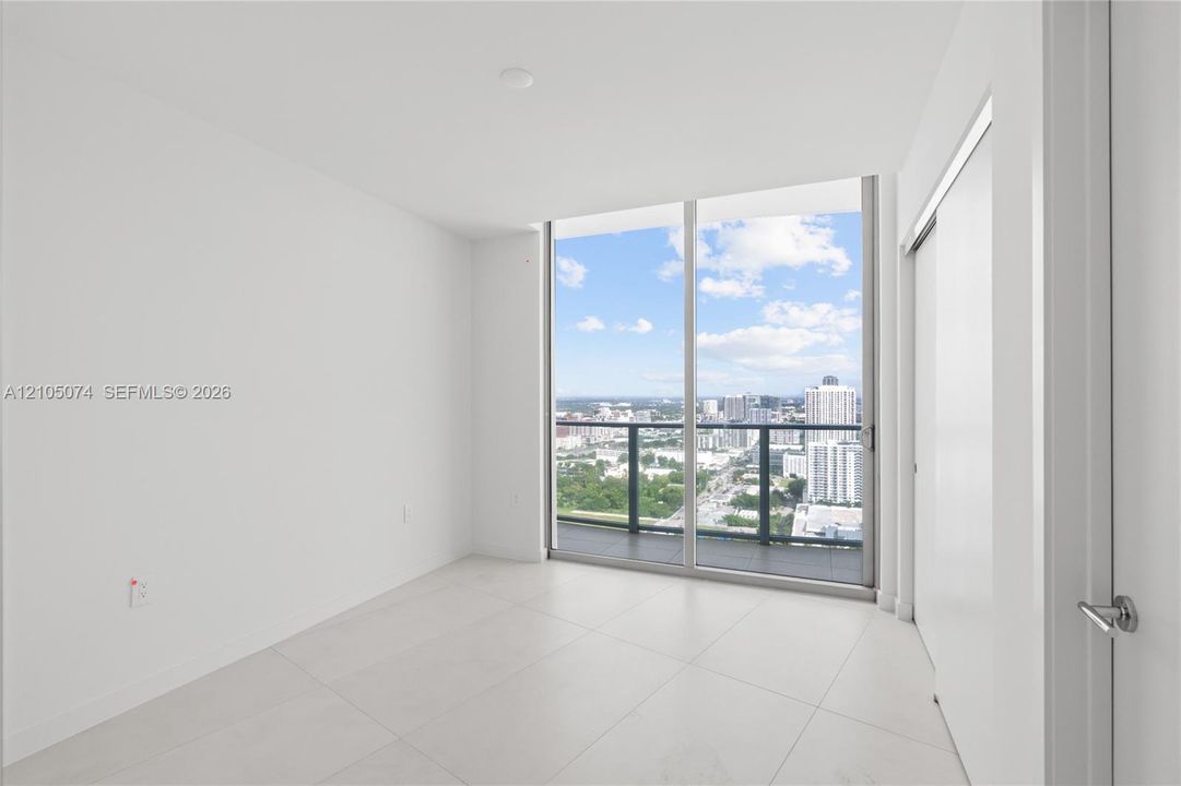 Floor-to-ceiling glass sliding doors, private balcony with metal railing, light-colored tile flooring, and urban horizon scenery