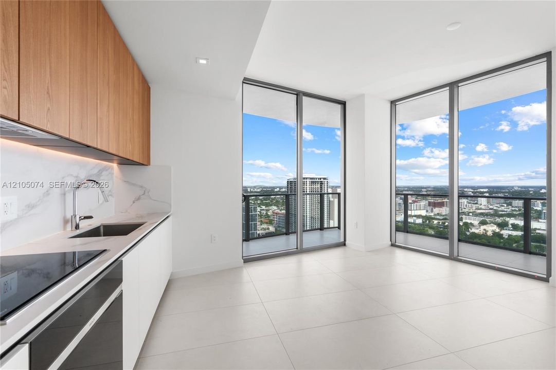 Modern kitchen featuring slab-front wood cabinetry, white stone-finish countertops, full-height stone-finish backsplash, built-in cooktop, and floor-to-ceiling windows with balcony access