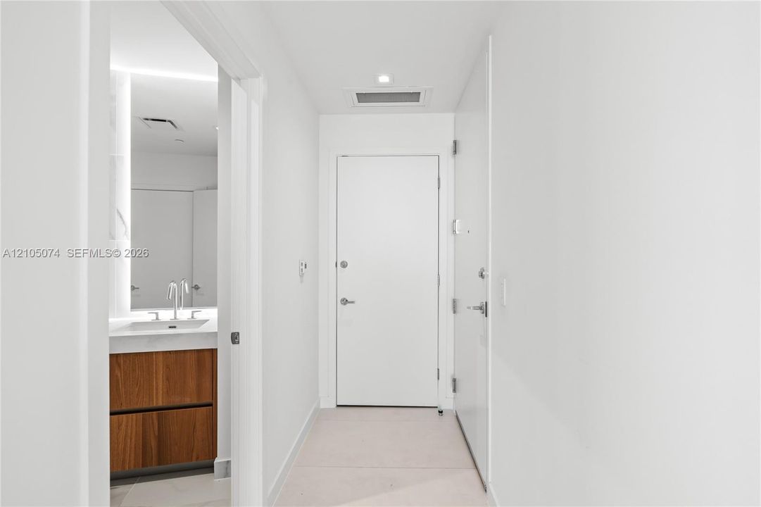 Neutral palette hallway featuring minimalist white interior doors, recessed lighting, and large-format tile flooring, with a glimpse of a wood-finish vanity and integrated basin