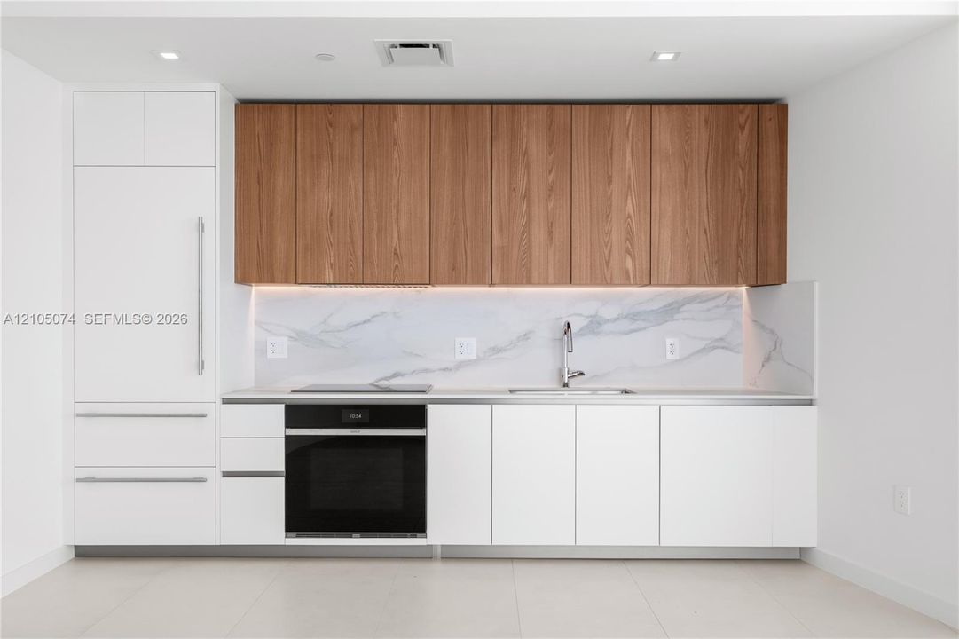 Modern kitchen featuring flat-panel white lower cabinetry, wood-finish upper cabinetry, integrated appliances, a stone-patterned backsplash, and recessed lighting