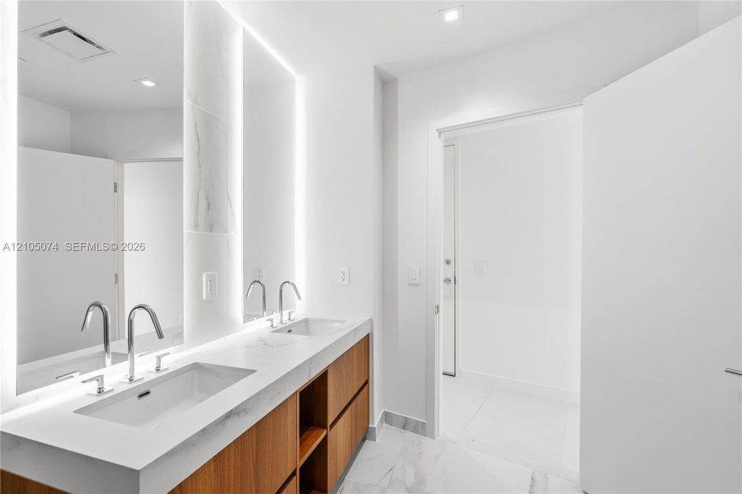 Floating wood-finish vanity with dual integrated sinks, light-veined stone countertop, back-lit vanity mirrors, and polished chrome fixtures