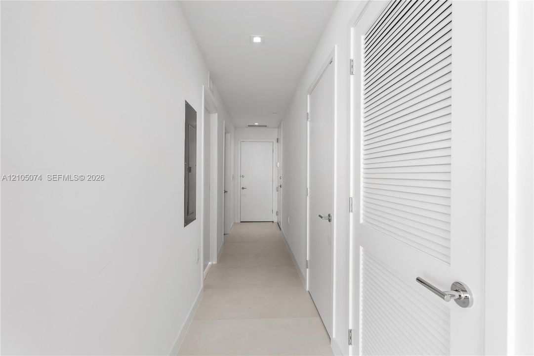 Hallway featuring neutral-toned tile flooring, recessed ceiling lighting, and solid core doors with brushed metal hardware, including a louvered closet door