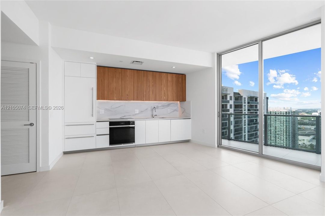 Kitchen featuring wood-finish cabinetry, white lower cabinets, stone-surfaced backsplash, integrated oven, and floor-to-ceiling glass doors leading to a private balcony