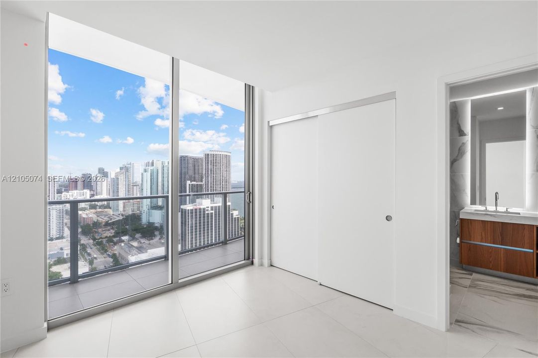 Floor-to-ceiling windows, private balcony with metal railing, sliding closet doors, wood-finish vanity with countertop basin, and stone-finish flooring