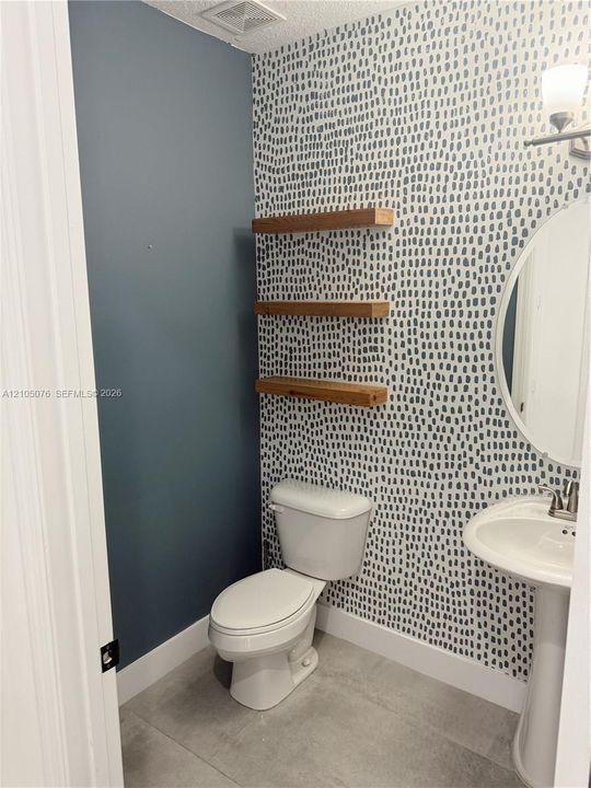 Patterned wallpapered accent wall, three built-in wood-finish floating shelves, white pedestal sink with metallic faucet, oval wall-mounted mirror, and tile flooring