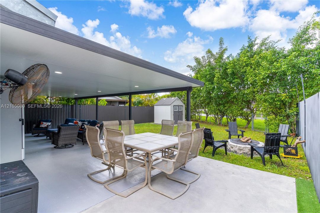 Covered outdoor patio featuring recessed lighting and a wall-mounted fan, stone-bordered fire pit, and enclosed backyard with perimeter fencing