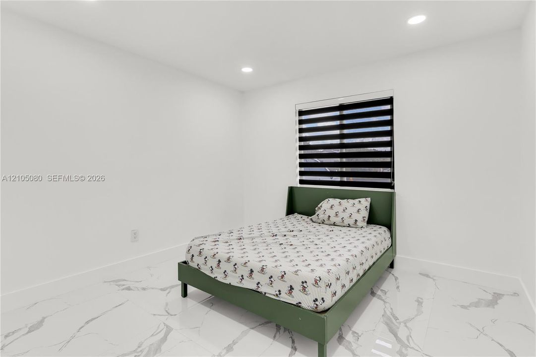 Bright room featuring white walls, white stone-finish tile flooring, recessed ceiling lighting, and a dual-shade roller window blind