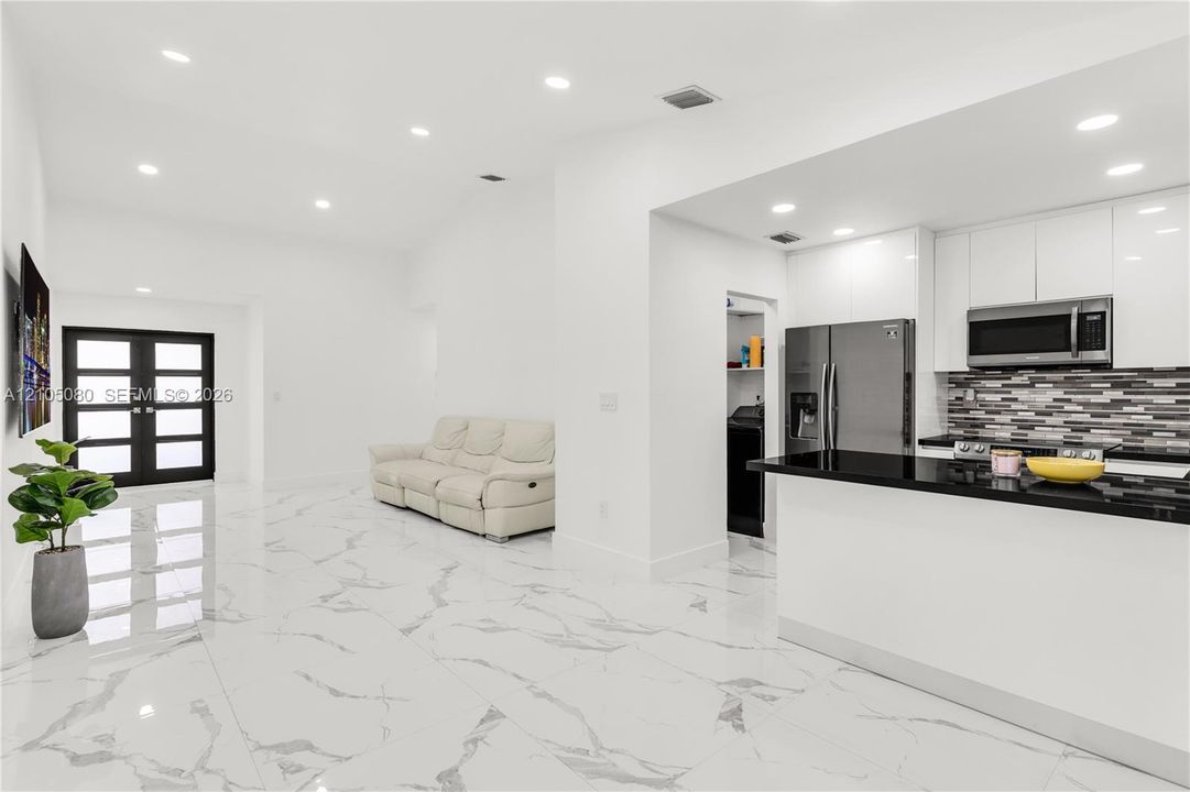 Interior space featuring high-gloss, large-format tile flooring, recessed ceiling lighting, a kitchen with white cabinetry and dark stone-surface countertops, a multi-pane glass-panel entry door, and a decorative tile backsplash
