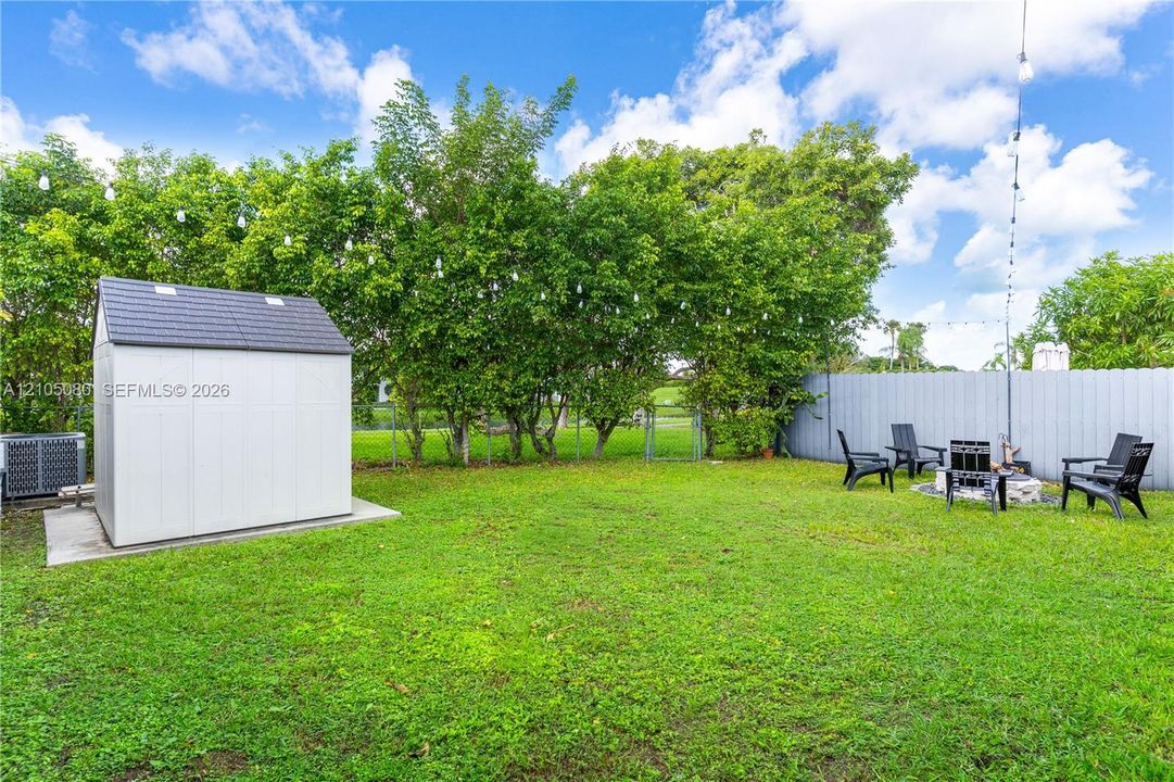 Fenced backyard featuring a manicured lawn, detached storage structure on a concrete pad, mature perimeter trees, and string lighting