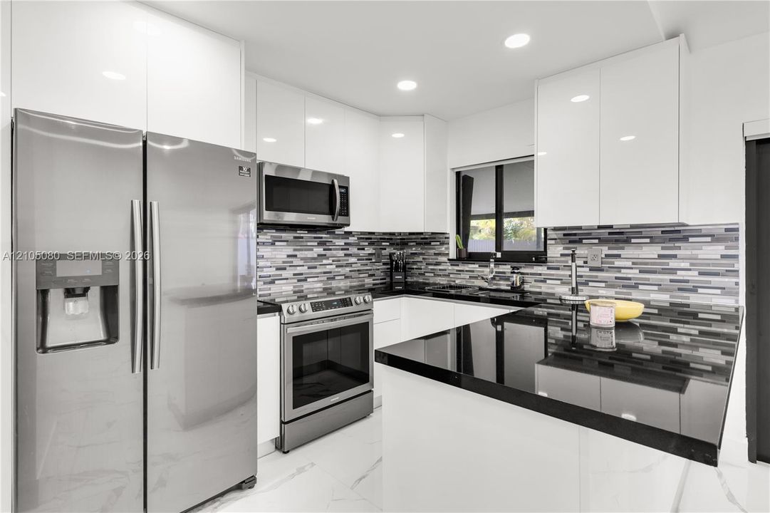 Kitchen featuring high-gloss white cabinetry, dark stone-finish countertops, stainless steel appliances, multi-toned mosaic tile backsplash, and large-format tile flooring