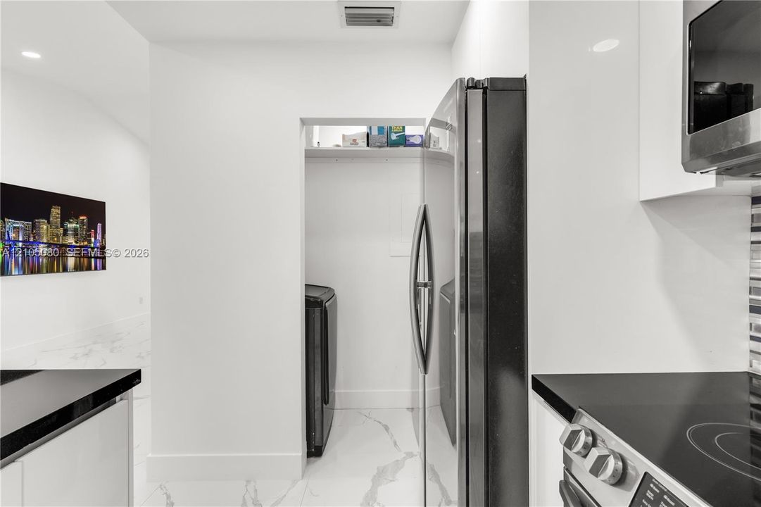 Kitchen featuring high-gloss white cabinetry, stone-finish tile flooring, a dark-toned integrated refrigerator, and a recessed laundry alcove with overhead shelving