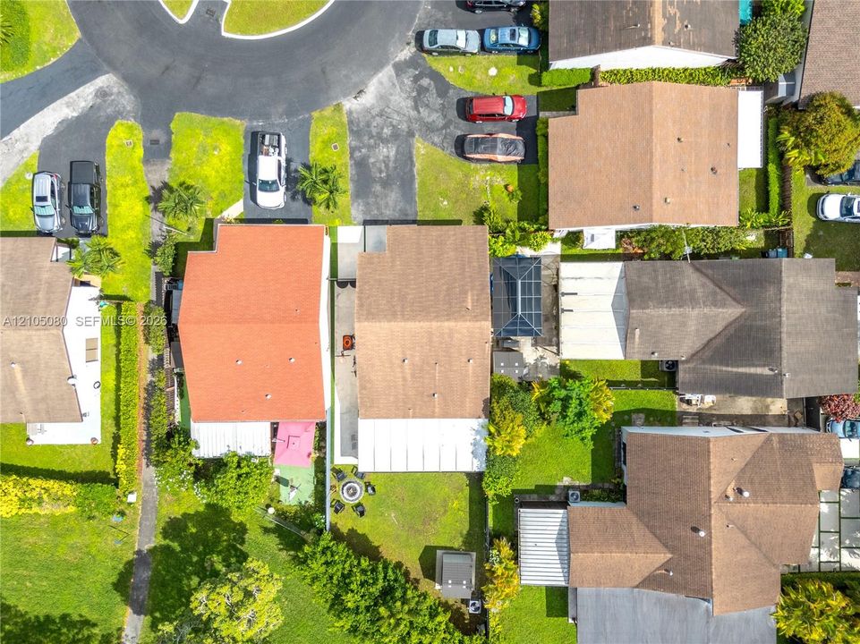 Aerial perspective of residential property featuring a multi-pitched shingle roof, paved driveway, enclosed patio structure, and established landscaping