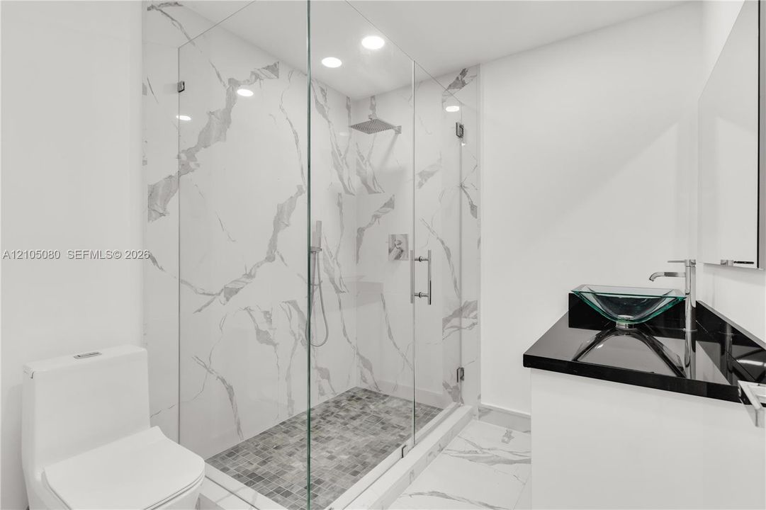 Walk-in shower with floor-to-ceiling veined tile surrounds and frameless glass enclosure, mosaic-tiled shower floor, recessed lighting, and vanity with dark countertop and vessel sink