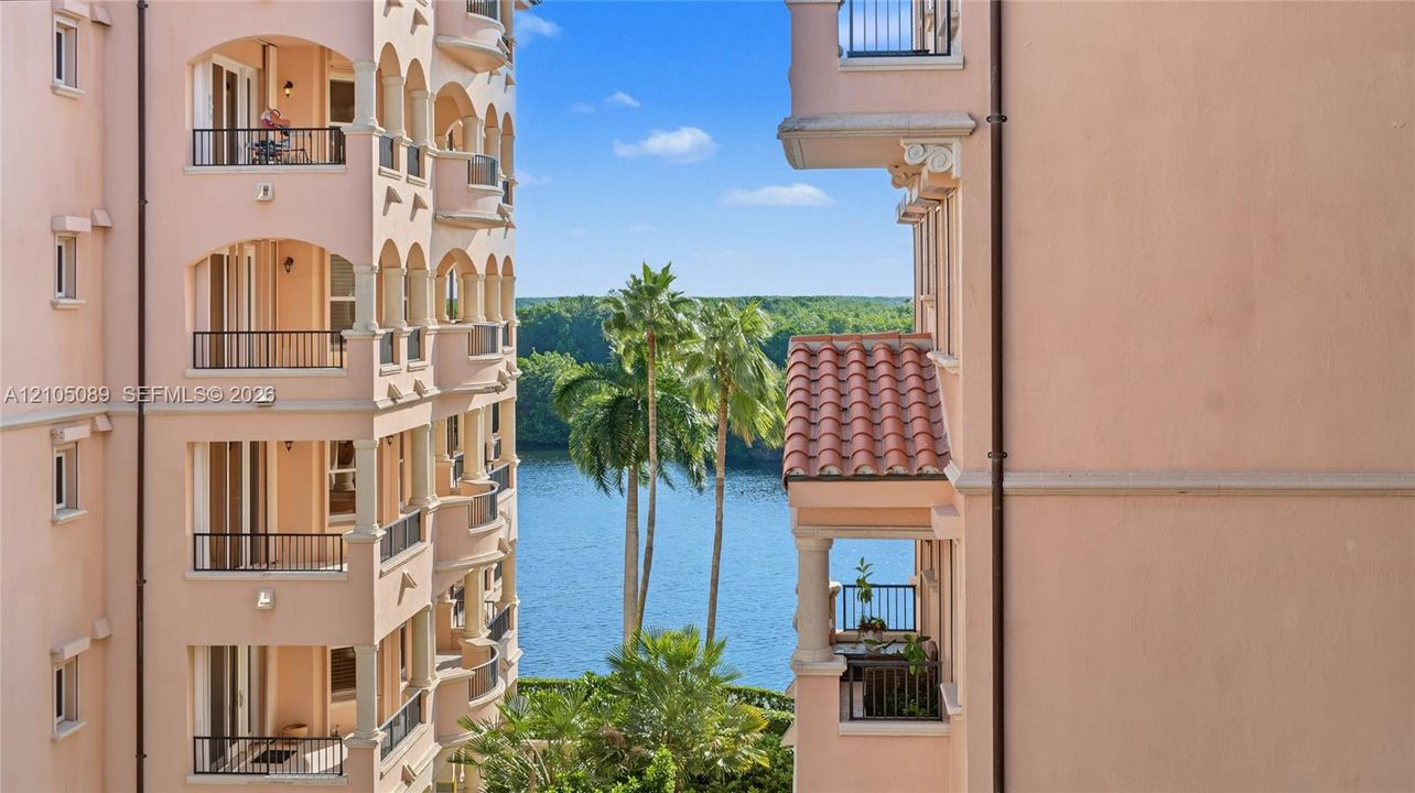Stucco exterior with multi-level arched balconies, metal railings, column supports, and clay tile roof accents, featuring a water backdrop with palm trees