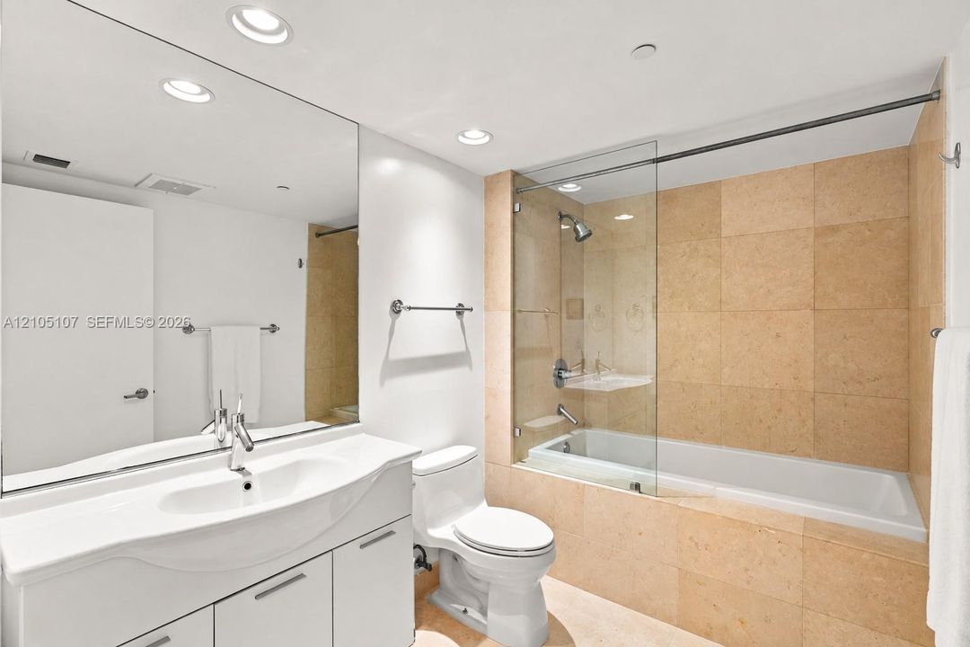 Full-length vanity mirror, white curved vanity with undermount sink, recessed lighting, and a bathtub with glass enclosure and tiled surround