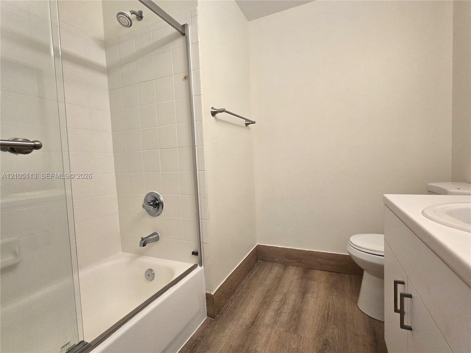 Bathtub with glass enclosure and shower head, white tiled shower surround, wood-finish flooring, white vanity with integrated sink, and white toilet