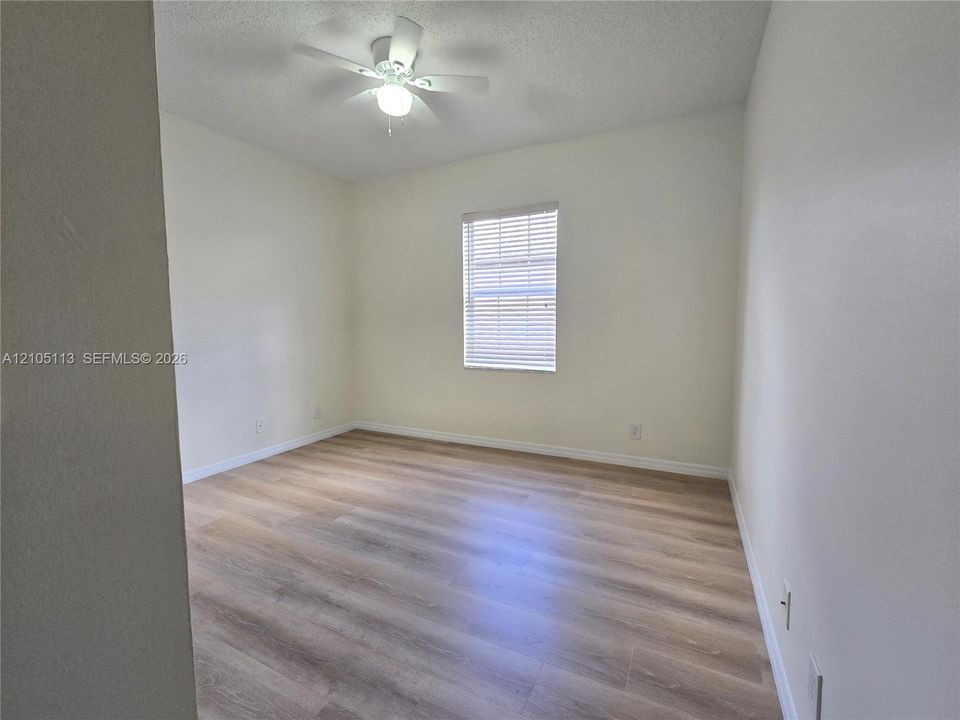 Bright, light-filled bedroom featuring wood-finish flooring, a ceiling fan with integrated lighting, and a large window with plantation shutters allowing natural illumination throughout the space