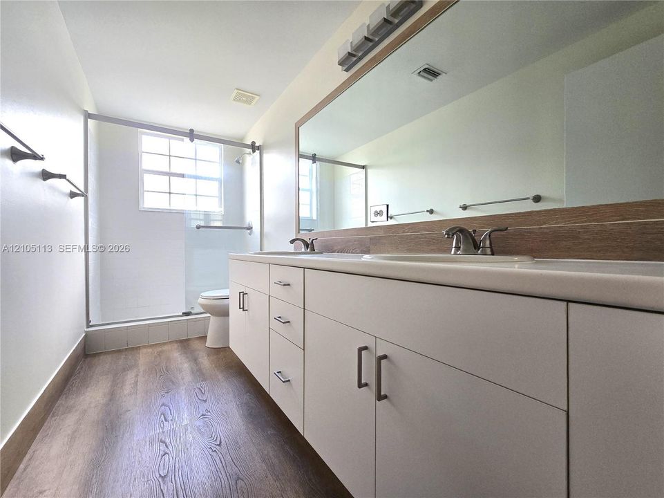 Dual vanity with integrated sinks and chrome fixtures, white cabinetry with dark pulls, full-width mirror with wood-finish trim, glass-enclosed shower, and wood-finish flooring