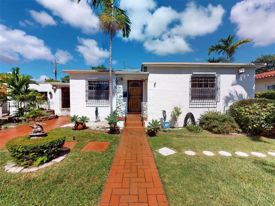 Single-story exterior featuring white stucco finish, red brick paver walkway, stone-accented entryway, and metal window security bars