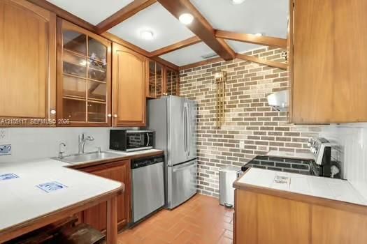 Kitchen featuring wood cabinetry with glass-front display panels, exposed ceiling beams, a decorative brick accent wall, tiled countertops, and tile flooring
