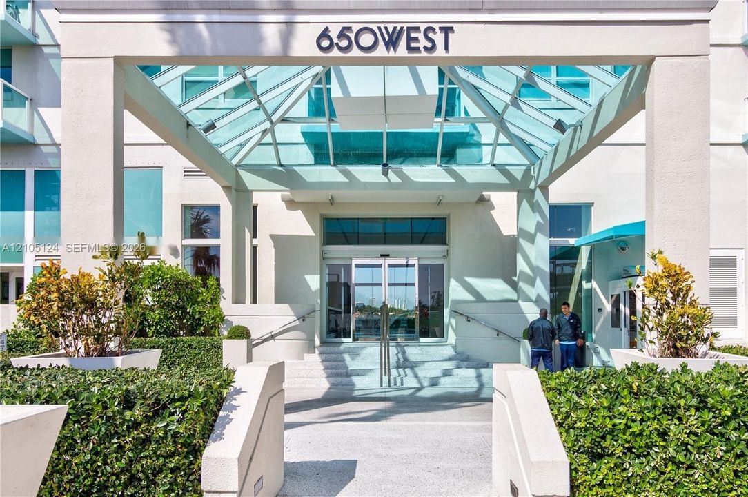 Contemporary building entrance featuring a glass canopy, automatic sliding doors, and integrated landscaping with structured hedges