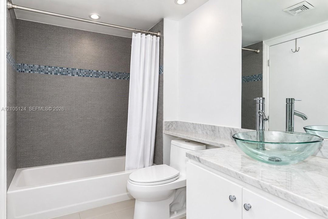 Spa-inspired bathroom featuring a white subway tile surround with decorative mosaic accent band, integrated soaking tub with white curtain rod and liner, vessel sink with polished chrome faucet mounted on marble-finish countertop, white cabinetry with hardware accents, and recessed ceiling lighting