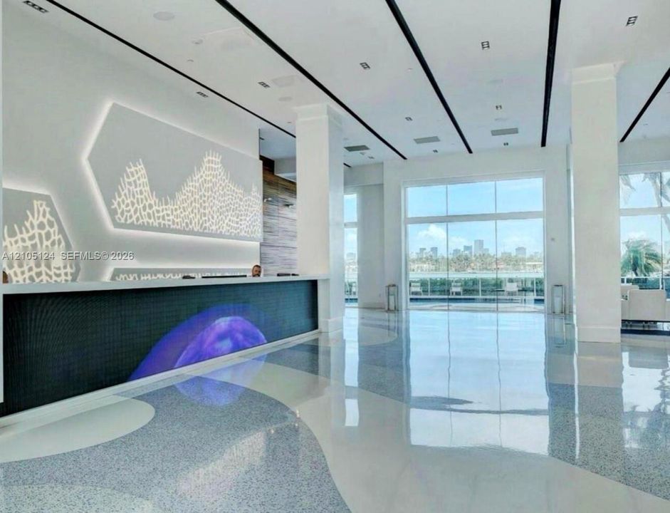Lobby area featuring large format windows, a patterned accent wall with integrated backlighting, a reception desk with a digital display, polished flooring, and structural columns