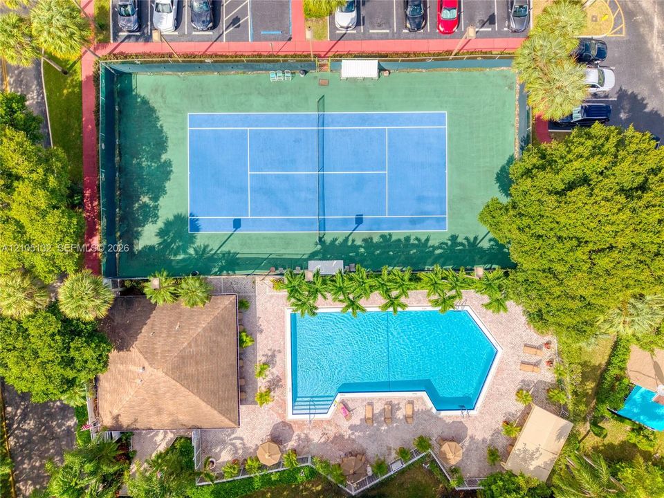 Outdoor tennis court with a blue playing surface, adjacent to a large swimming pool with a paved surround, and a nearby building featuring a brown shingle roof
