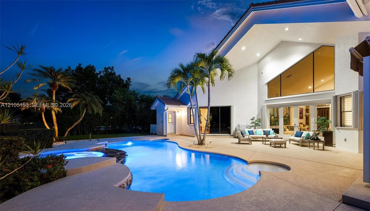 Curved swimming pool with integrated spa and tiled accents, expansive patio with concrete surfacing, and tropical landscaping