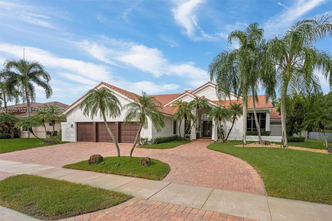 White stucco exterior with a terra cotta tile roof, a curved paver driveway, a two-car garage with wood-look doors, and lush landscaping