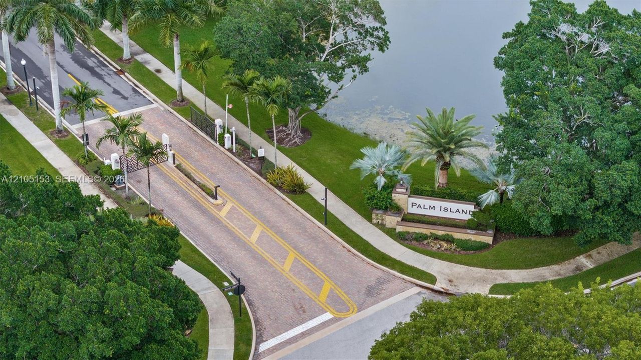 Community entrance with "Palm Island" signage, gated access, mature palm trees, and water feature